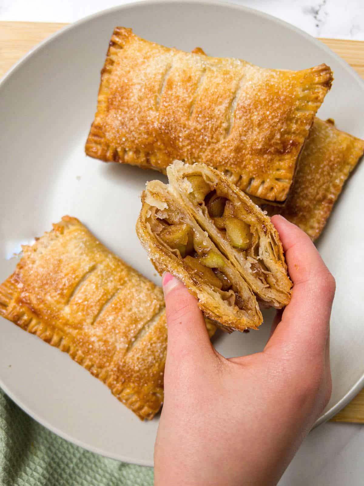Three apple turnovers on a neutral plate and one cut in half being picked up.