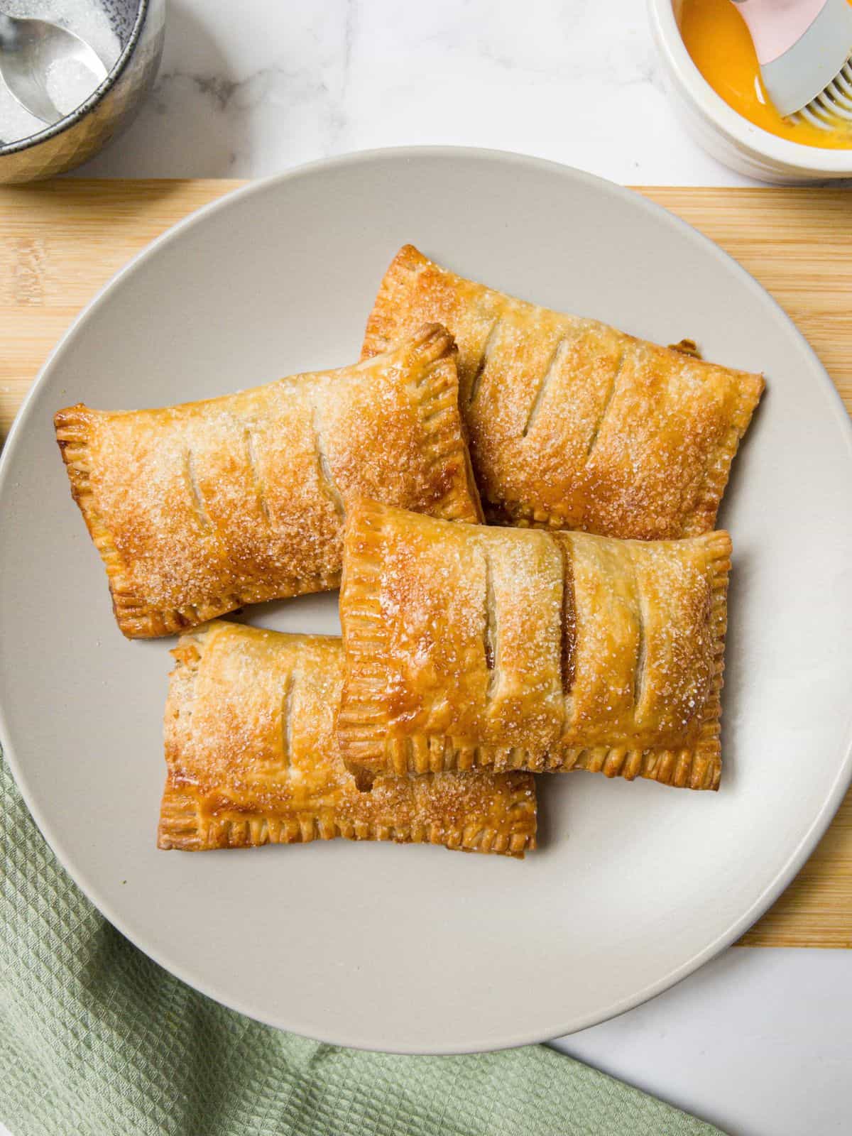 Four gluten free apple turnovers on a neutral coloured plate sat on a wooden chopping board.