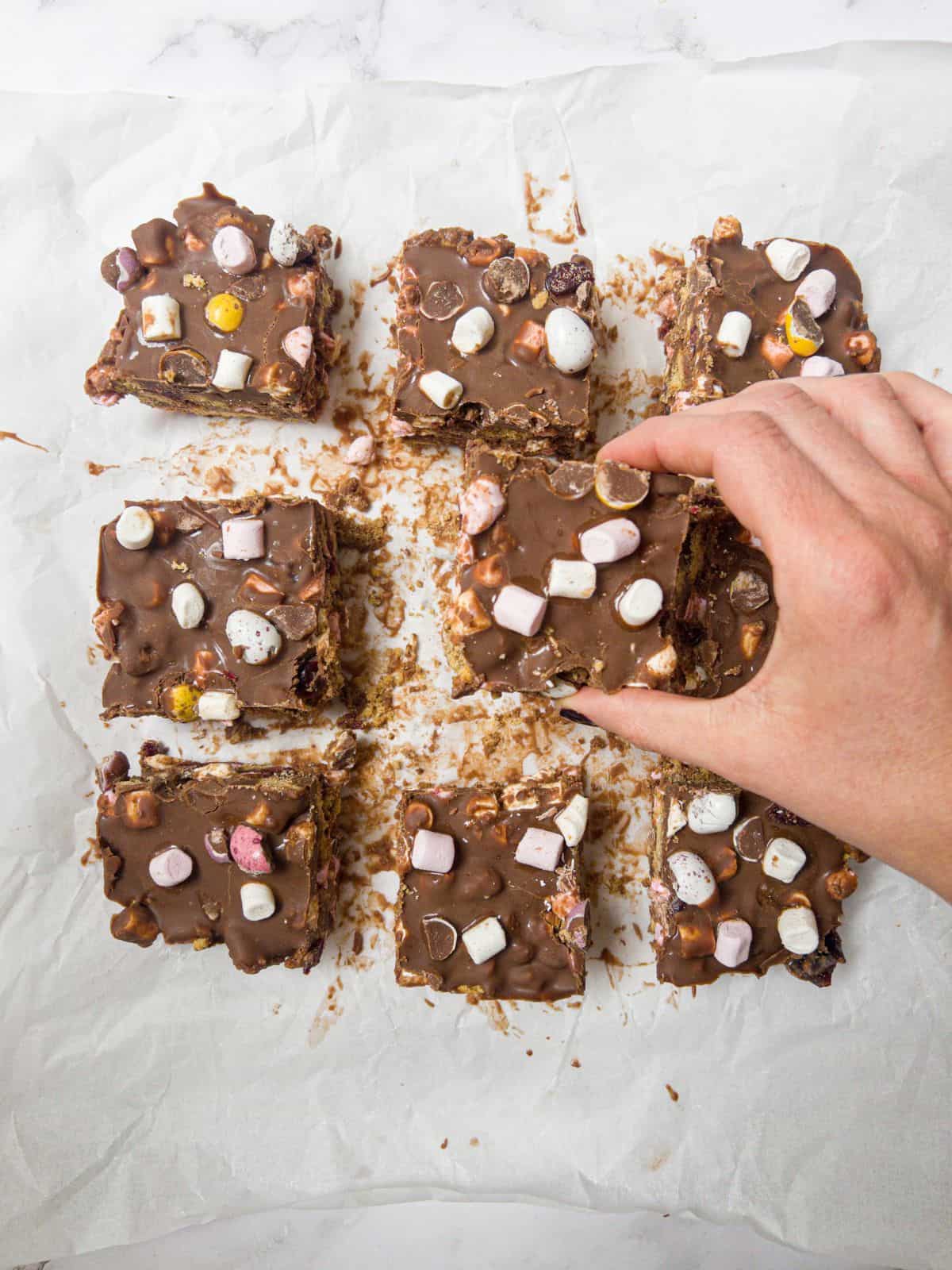 Nine cut squares of rocky roads sitting on greaseproof paper, with one slice being picked up by hand.
