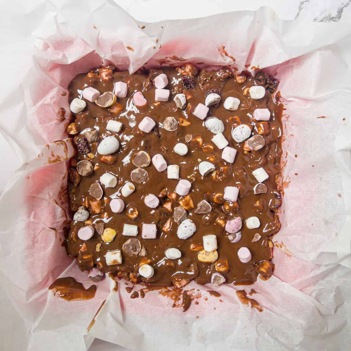 Un-set rocky road mix topped with marshmallows and mini eggs in a greaseproof paper lined baking tin.