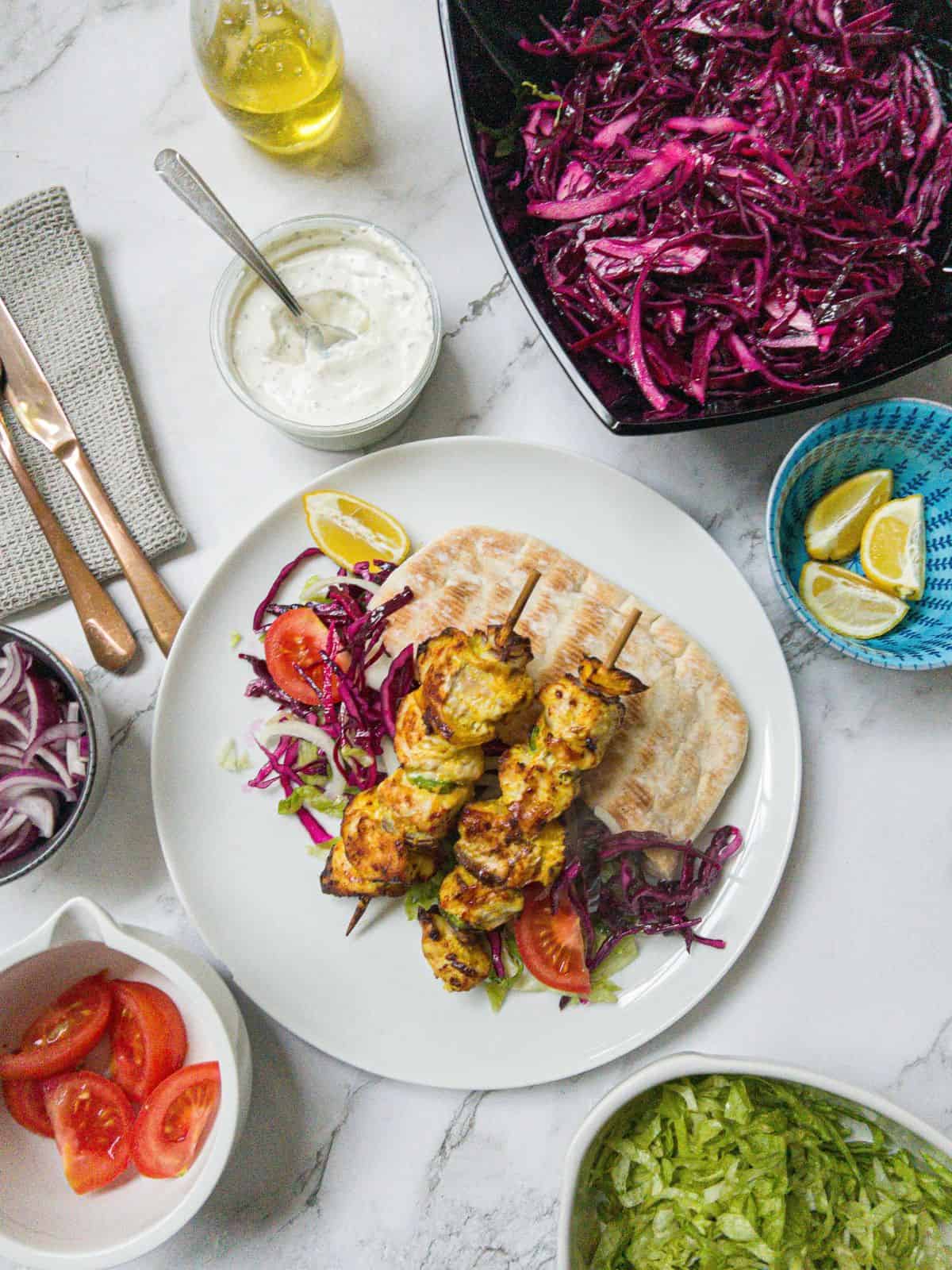 Two chicken shish kebabs on sticks laying on top of salad and pitta bread on a white plate. With a bowl of red cabbage, a pot of garlic sauce, lettuce and tomato in the background.