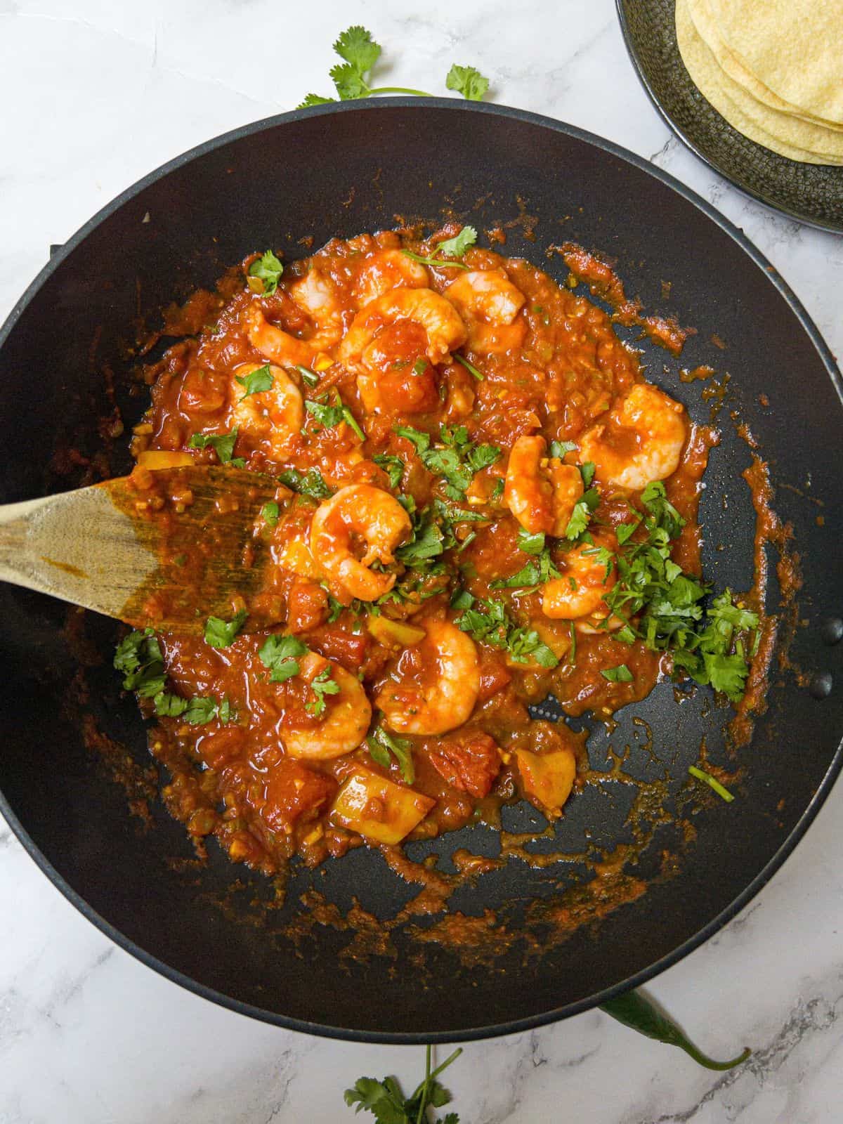 Prawn balti garnished with coriander in a black frying pan being stirred with a wooden spoon.