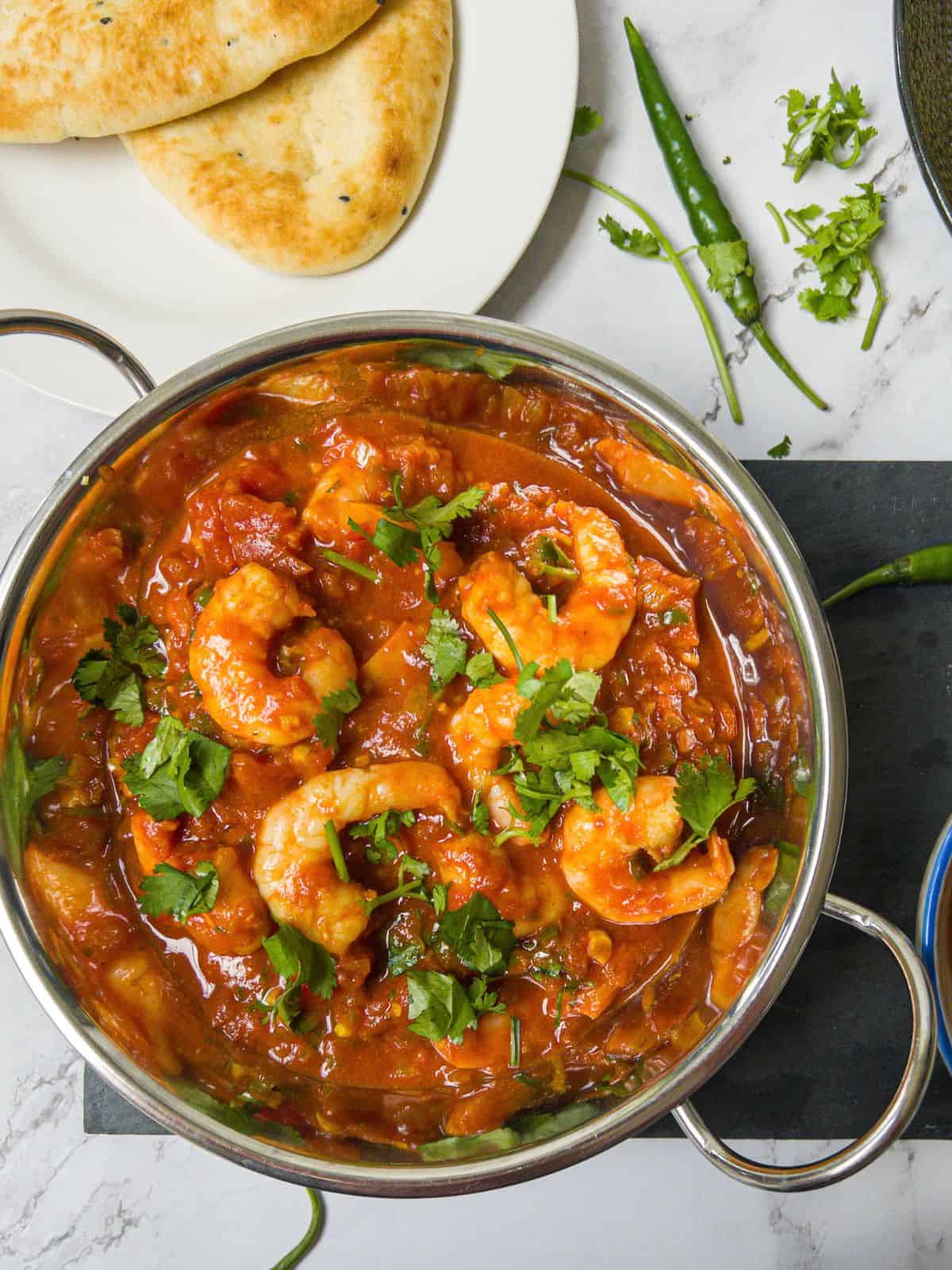 Prawn balti in a stainless steel bowl with naan bread and green chillis in the background.