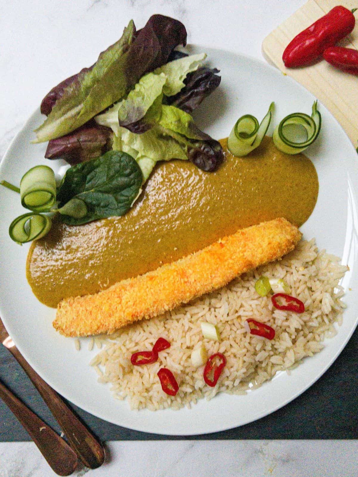 A white plate with a breaded salmon fillet, katsu curry sauce, rice and salad. Garnished with chillis.