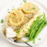 A plate containing halibut garnished with lemon on a bed of mash with green beans.