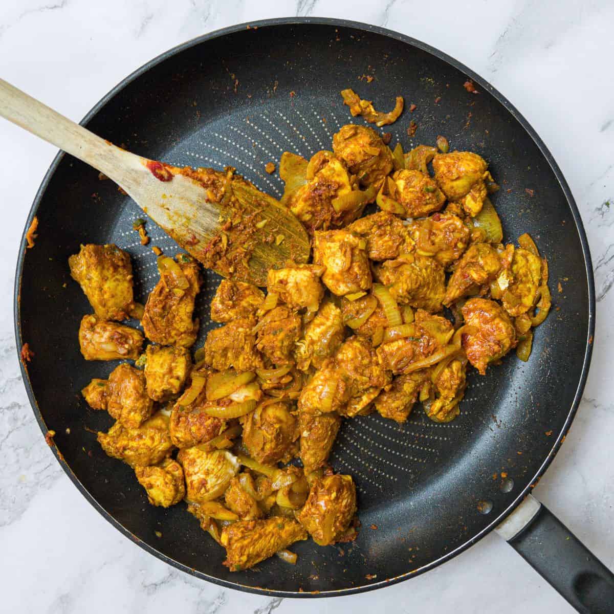 Chicken chunks and onions in a curry paste in a frying pan being stirred with a wooden spoon.