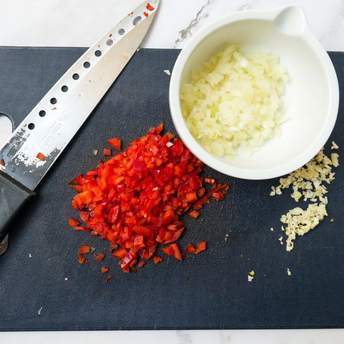 Chopped chilli, onion and garlic on a blue chopping board. A knife is also on the chopping board.