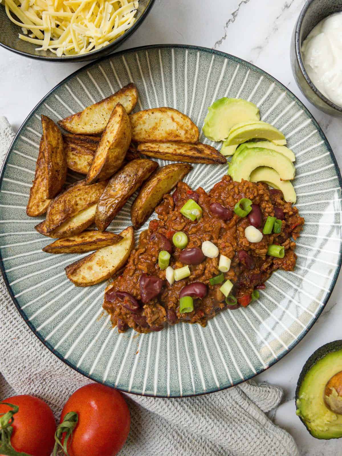 A blue plate containing chilli con carne, potato wedges and avocado. In the background are some tomatoes, avocado, a pot of cheese and a pot of sour cream.