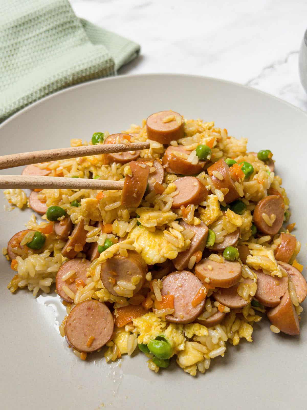 Hot dog fried rice with egg and peas on a plate with chopsticks pickling up some hotdog.