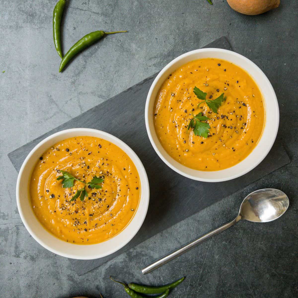 Two white bowls containing sweet potato, coconut & chilli soup. A spoonful is being taken out of one of the bowls.