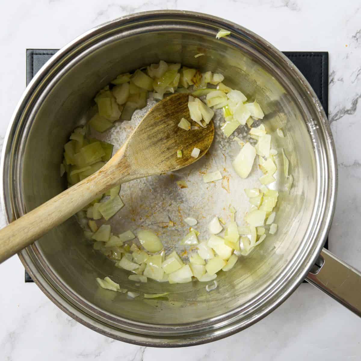 Diced onion frying in a saucepan, being stirred with a wooden spoon.