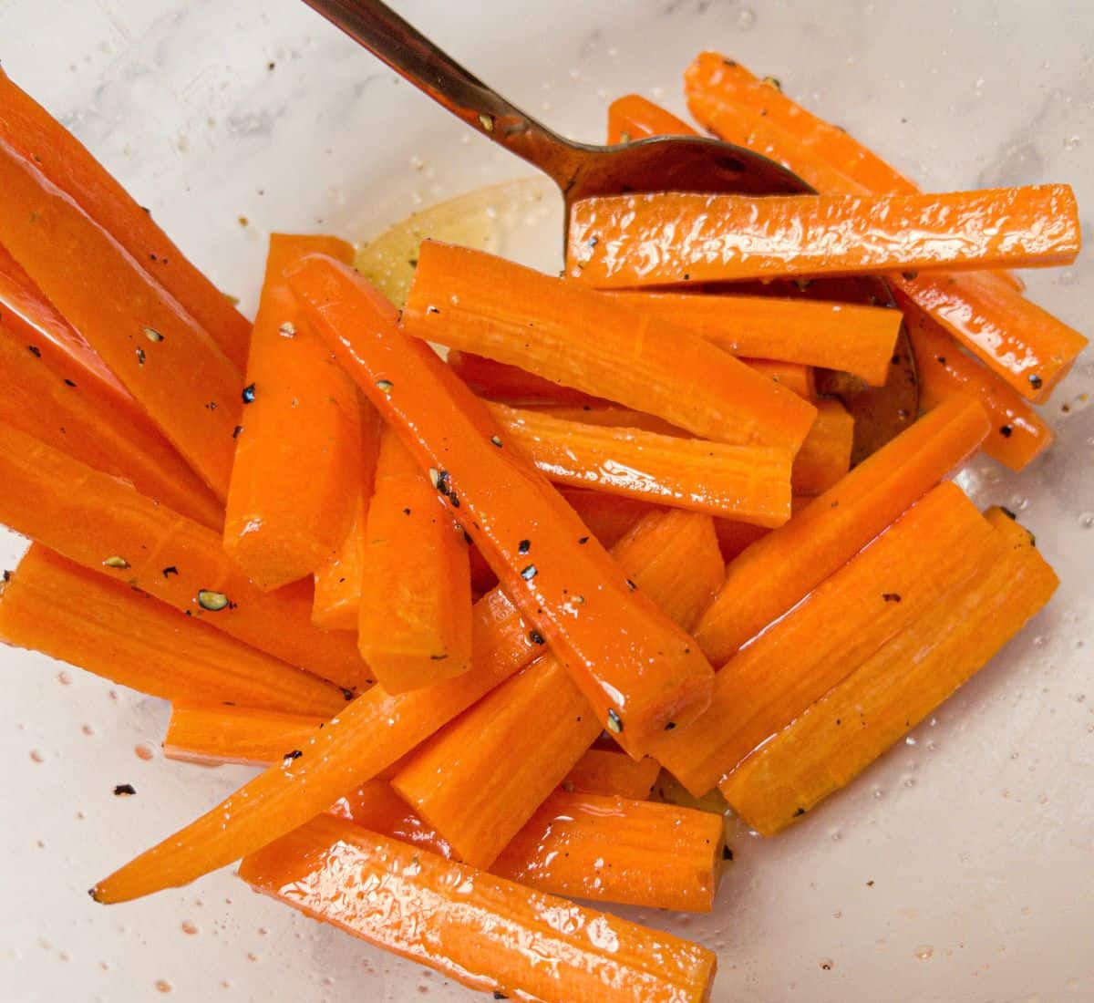 Carrot sticks tossed in oil and honey being mixed with a spoon in a bowl.
