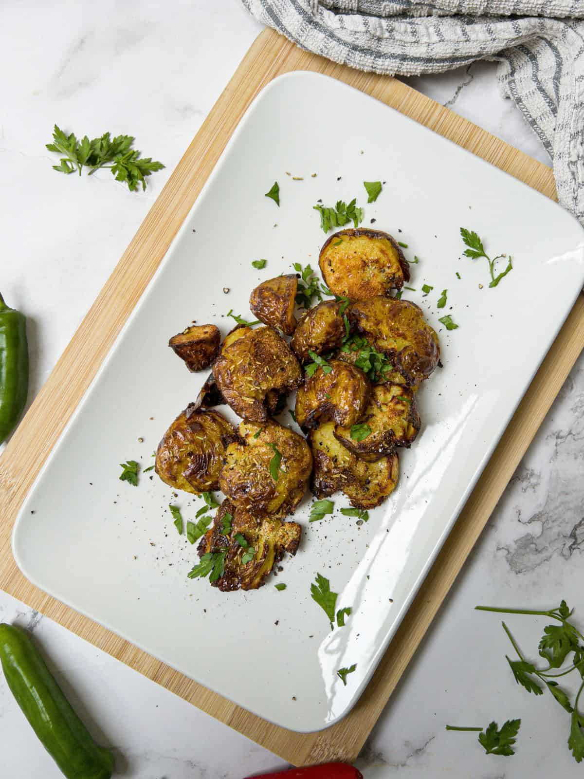 crushed new potatoes on a rectangle white plate garnished with parsley. Green chillies and parsley in the background.