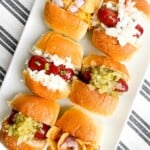 6 mini hots dogs with different toppings on a white oblong plate.