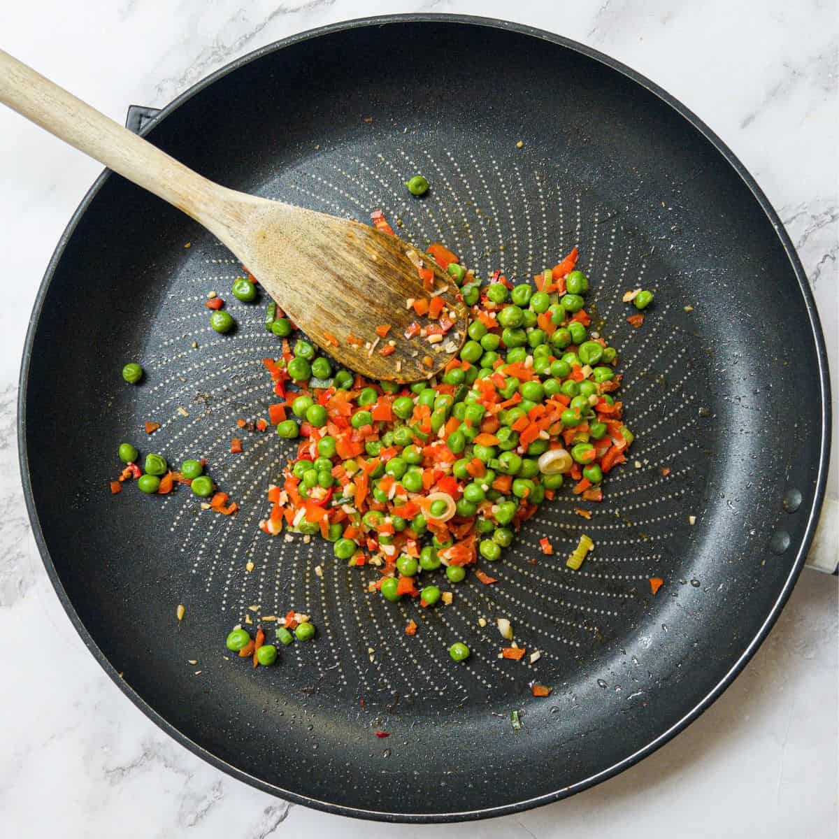 Peas and red peppers in a frying pan with a wooden spoon.