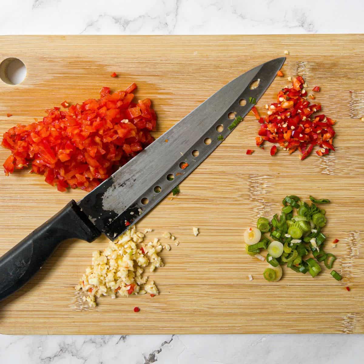 Red pepper, chilli, garlic & spring onion on chopping board with a knife.