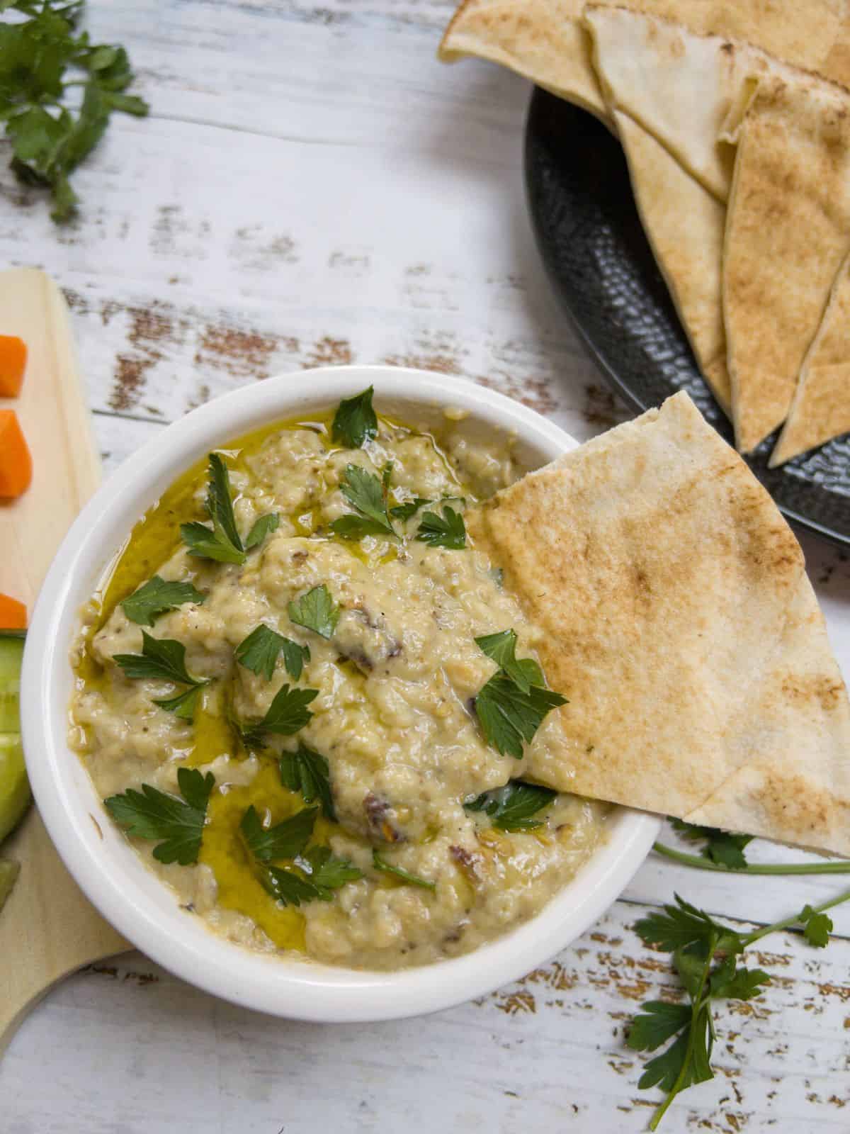A bowl of baba ganoush with a triangle of pitta bread in it. A plate of pitta sits in the background.