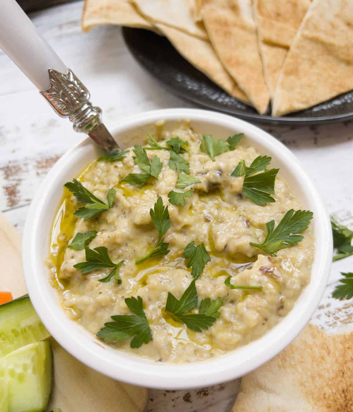 a bowl of baba ganoush garnished with parsley. Triangles of pitta bread in the background.