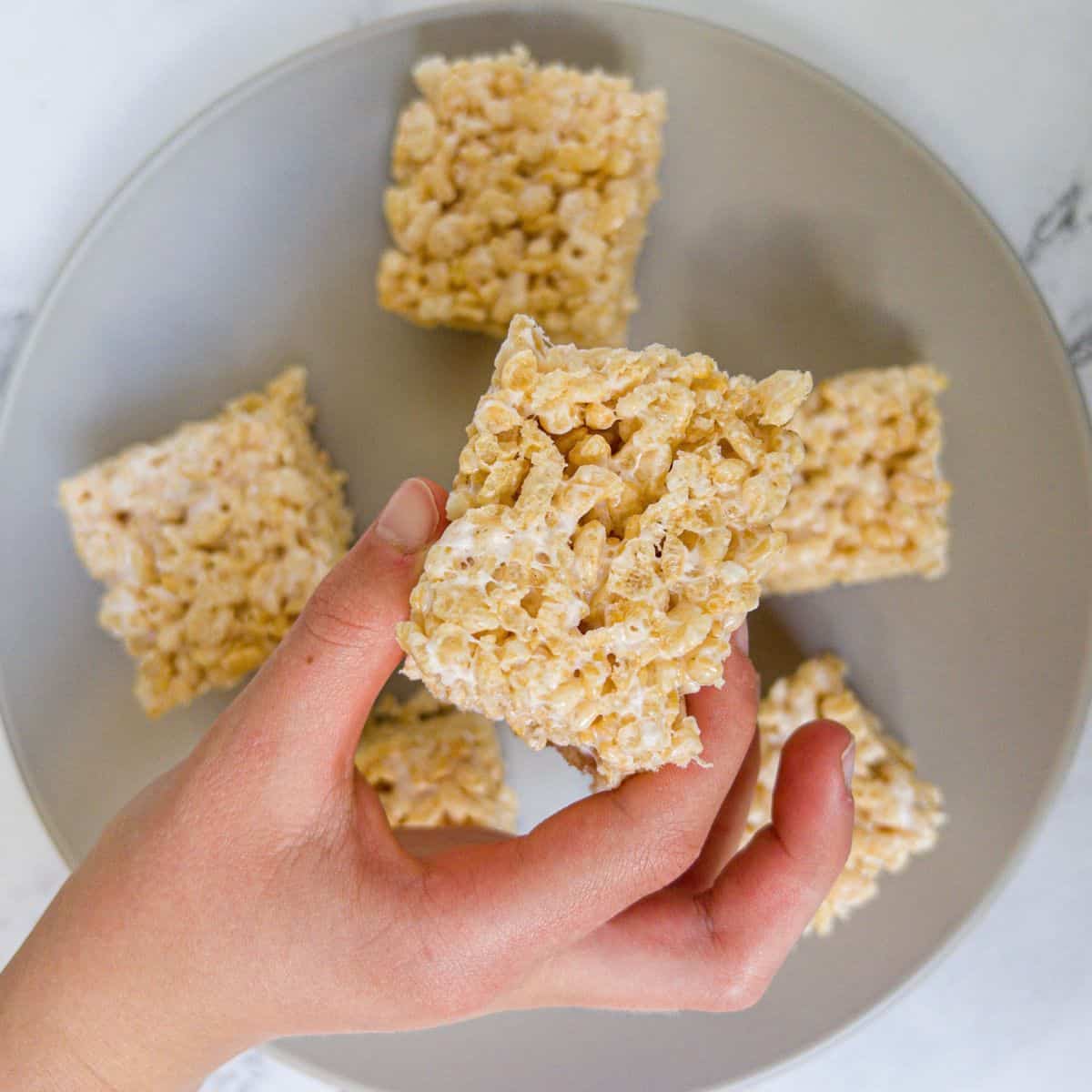 Squares of rice krispies mixed with melted marshmallows on a plate, one is being picked up.