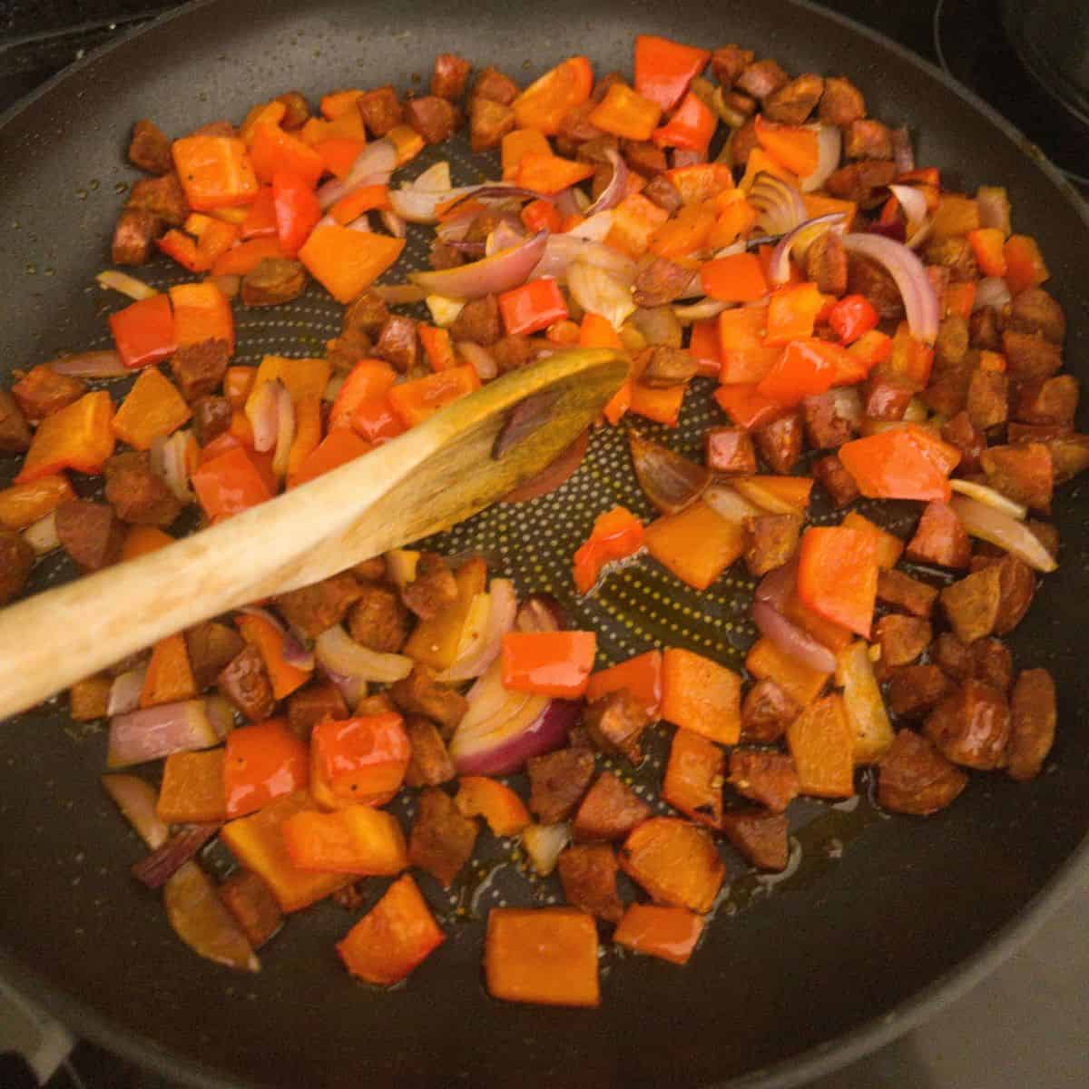 Chorizo, chunks of red pepper and red onion being stirred in a frying pan.