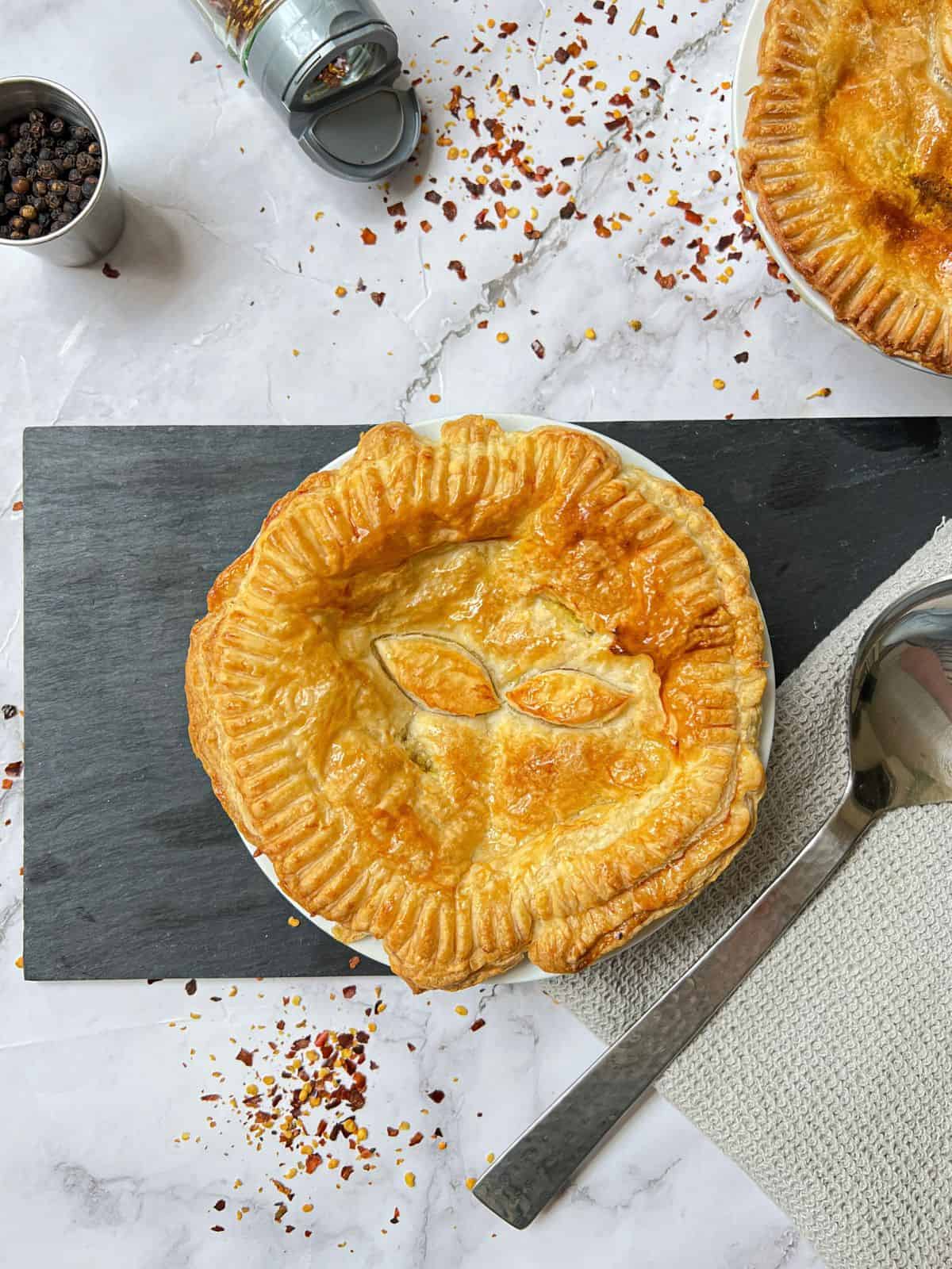 A pie garnished with leaf decorations with another pie and spilt chilli flakes in the background.