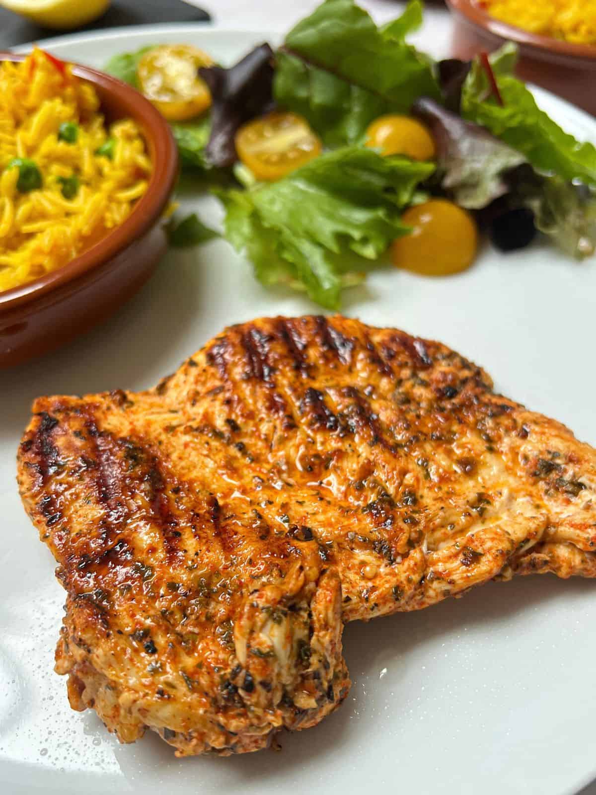 Griddled chicken breast on a plate with a bowl of rice and salad.