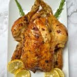 A roast chicken on a white plate garnished with rosemary and lemon.