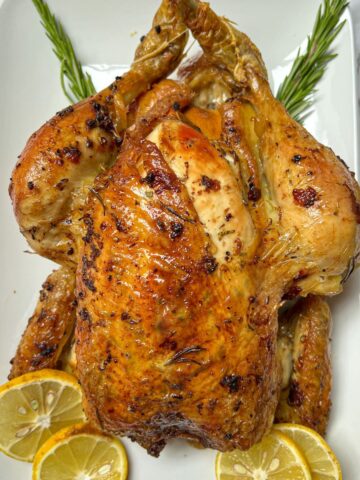 A roast chicken on a white plate garnished with rosemary and lemon.
