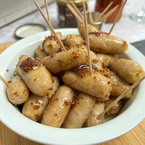 A white bowl containing honey and mustard coated cocktail sausages, with cocktail sticks poking out of them.