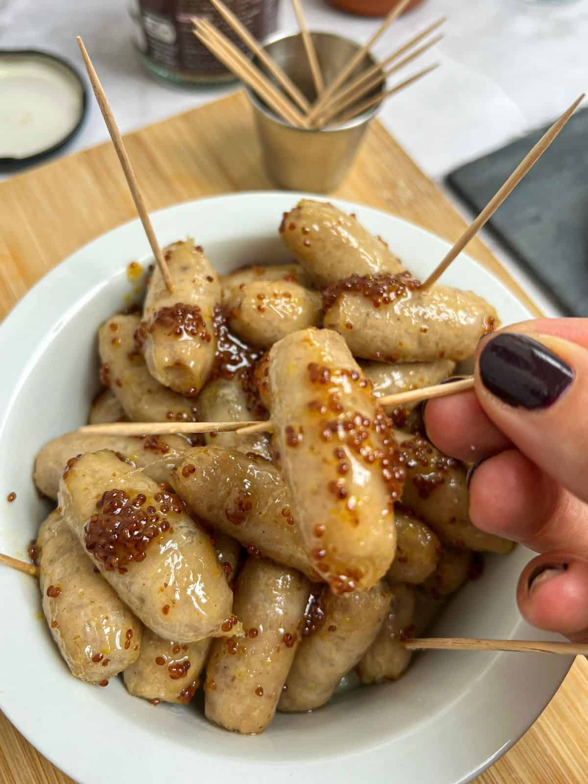 A white bowl containing honey and mustard-coated cocktail sausages, with one being picked up with a cocktail stick.