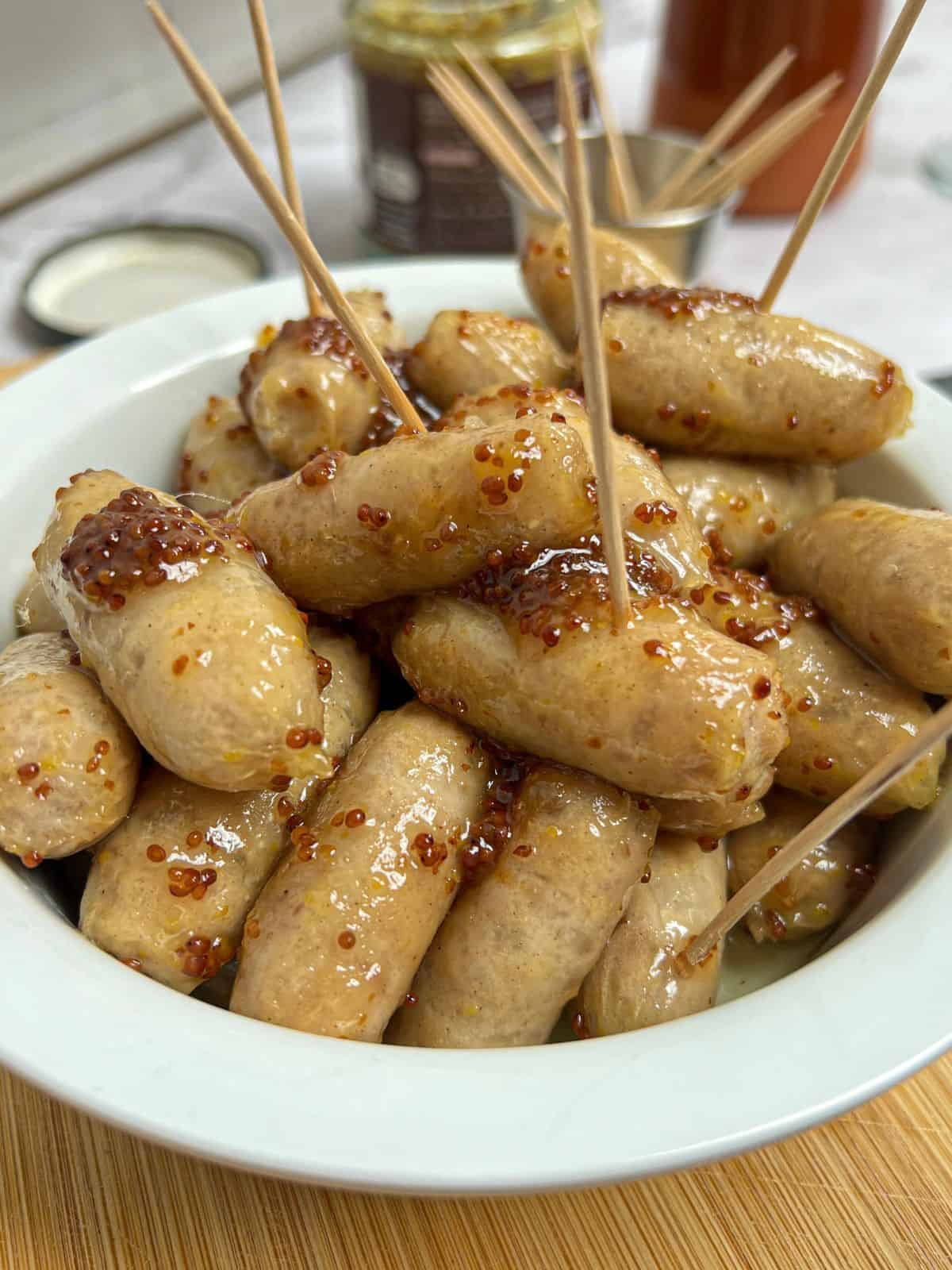 A white bowl containing honey and mustard-coated cocktail sausages, with cocktail sticks poking out of them.