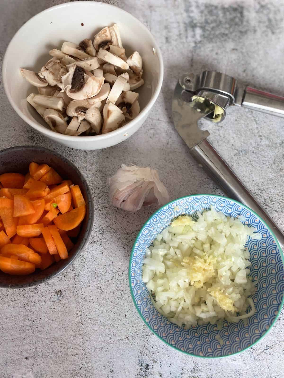 Three bowls containing chopped mushrooms, carrots and onions. Placed next to them is a garlic press and a bulb of garlic.