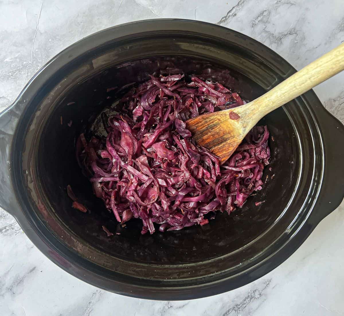 Cooked red cabbage in a slow cooker with a wooden spoon in it.