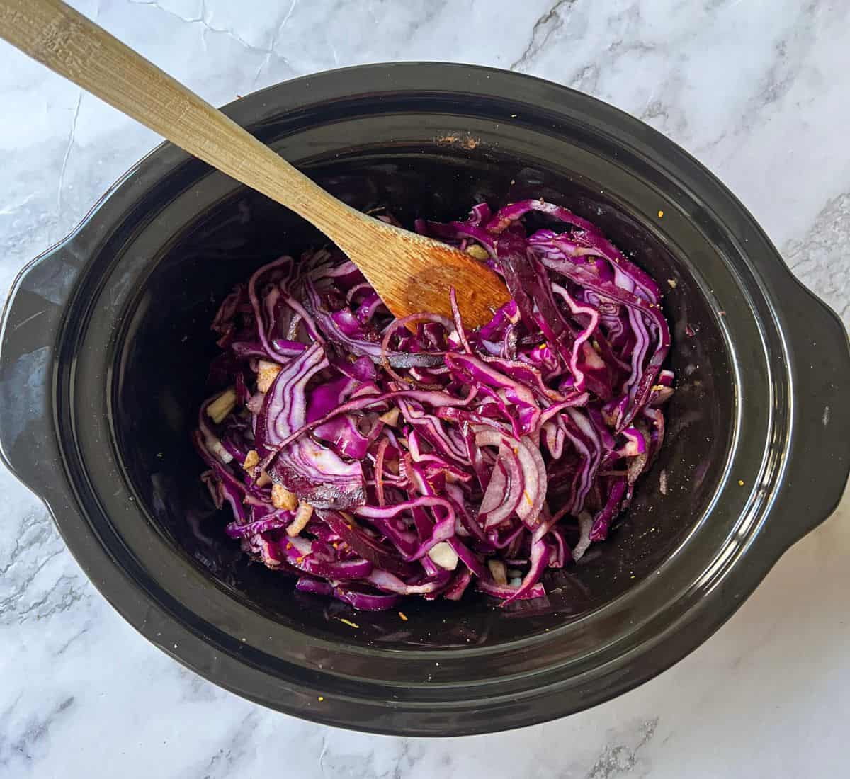 Sliced red cabbage in a slow cooker with a wooden spoon in it.
