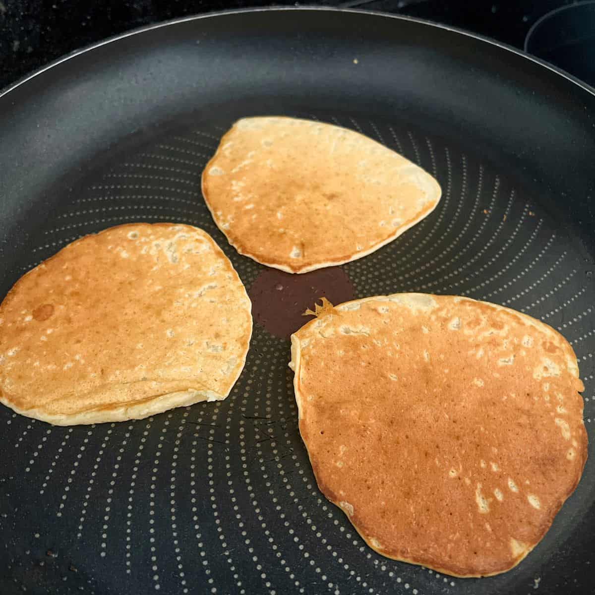 Three pancakes cooking in a frying pan.