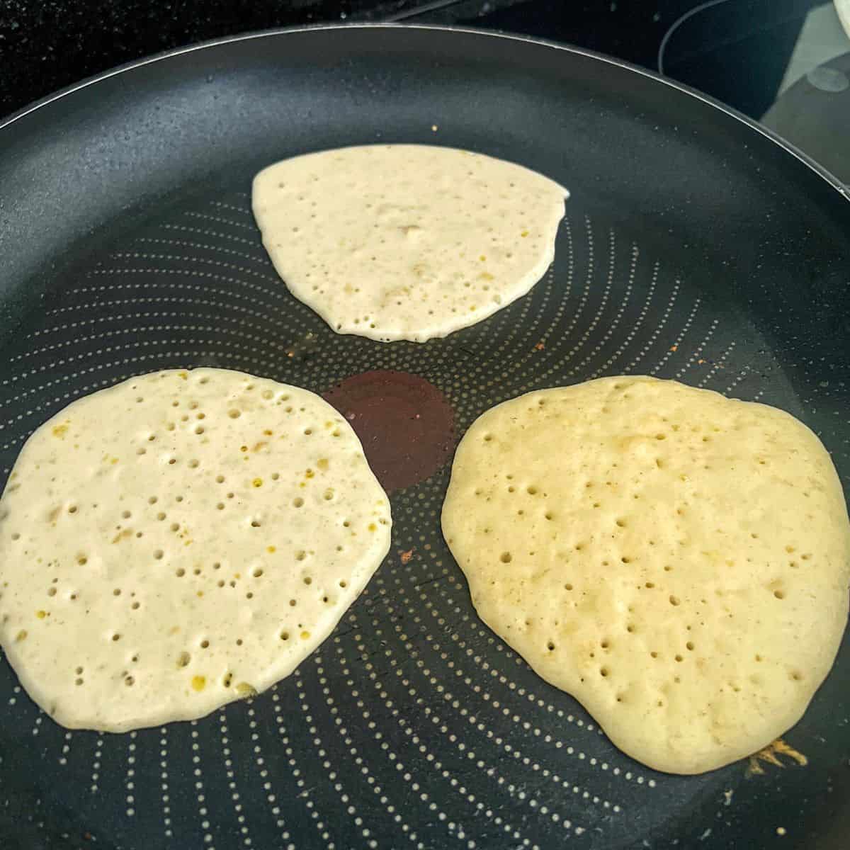 Three pancakes cooking in a frying pan.