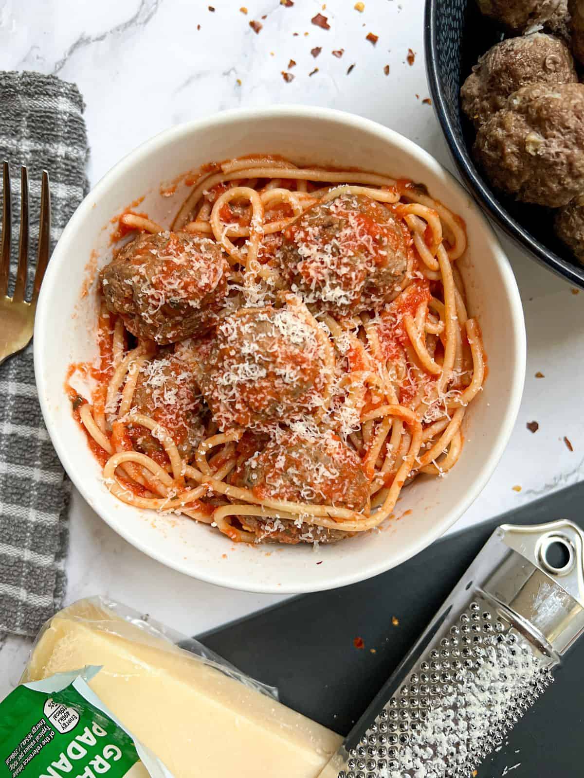 A bowl of spaghetti and meatballs with parmesan and a grater in the background.