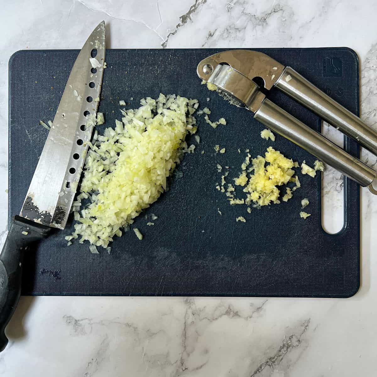 Chopped onion and garlic on a chopping board with a knife and garlic press.