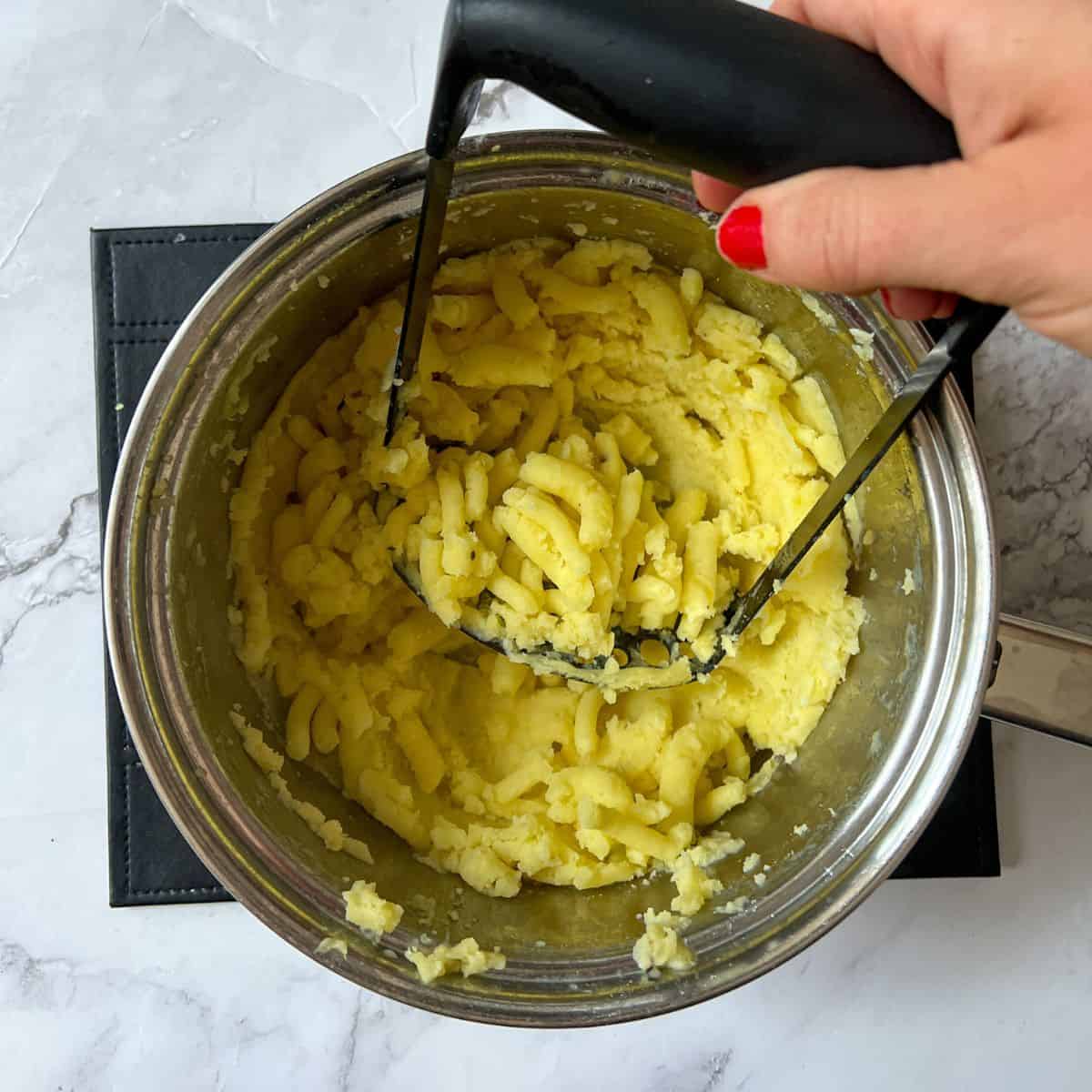 Potato in a saucepan being mashed with a potato masher.
