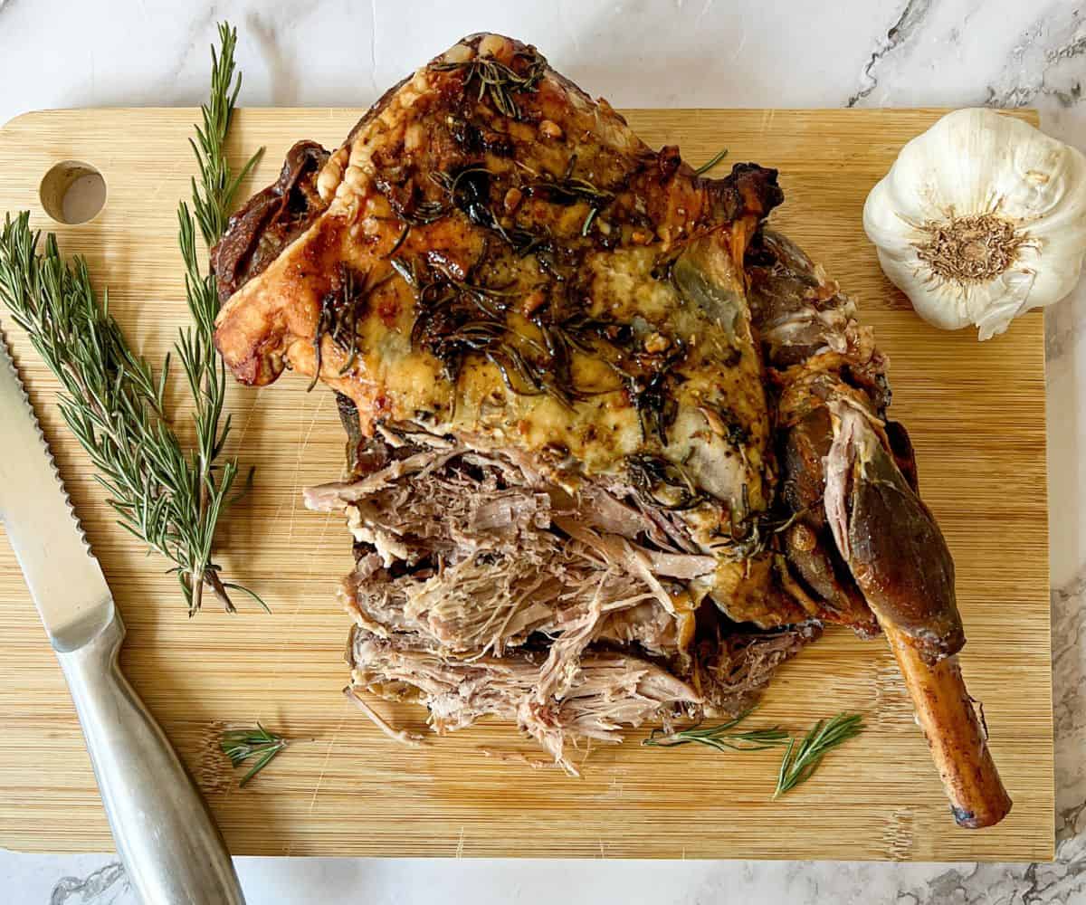A carved leg of lamb on a chopping board with a knife. There is a bulb of garlic and a sprig of rosemary also on the chopping board.
