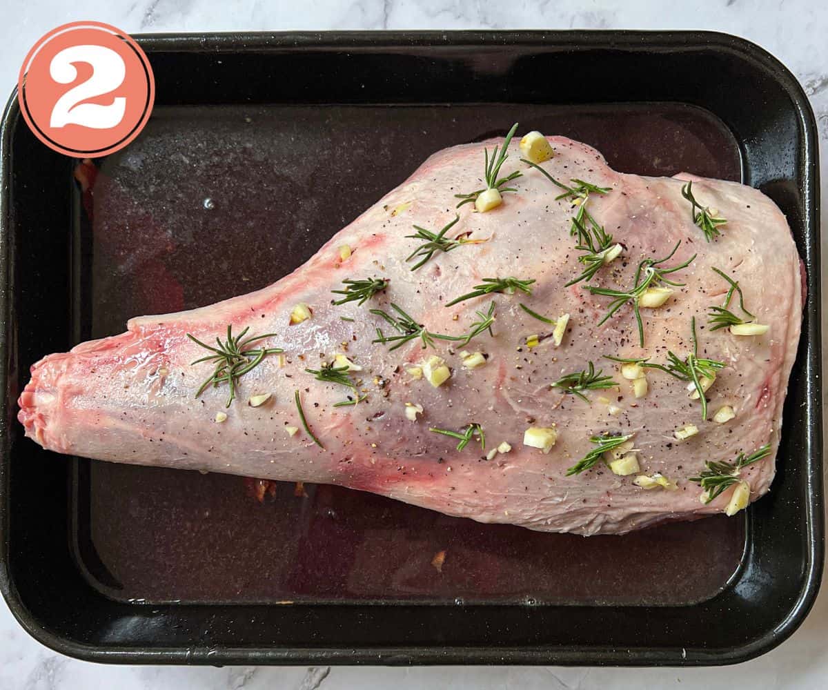 A raw leg of lamb studded with garlic and rosemary, sitting in a roasting tin.