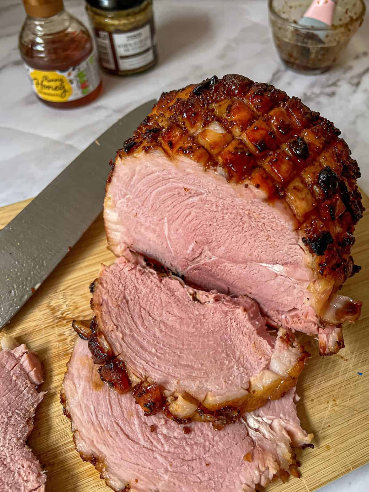 Carved ham (gammon) on a chopping board that has a knife on. A bottle of honey and a jar of mustard are in the background.