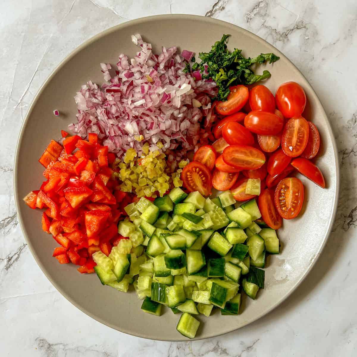 a plate containing chopped red pepper, cucumber, red onion, basil and chopped tomatoes.