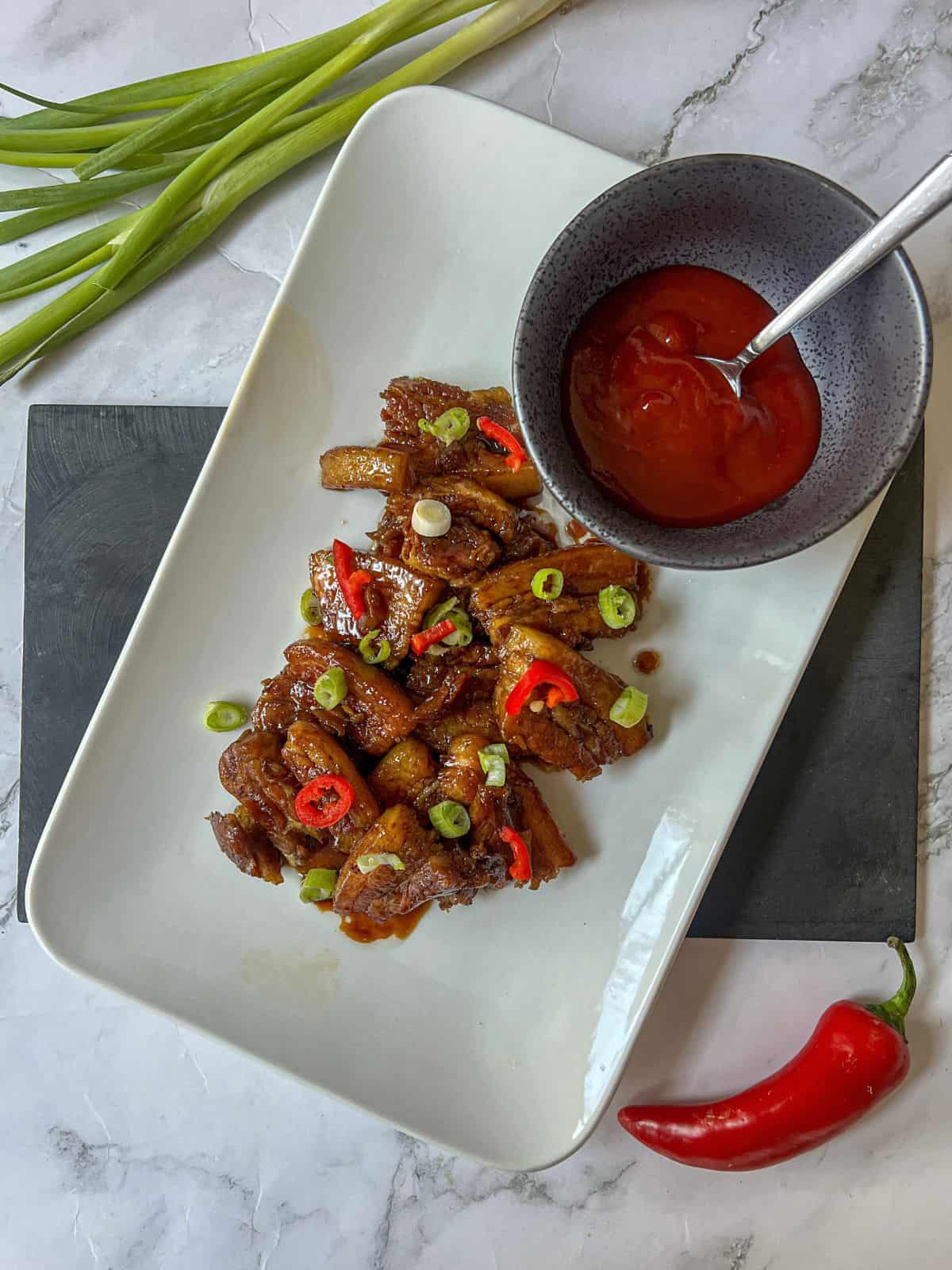 Chopped up pork belly slices in a sauce garnished with spring onion and chilli on a white plate. A bowl of dipping sauce is on the plate.