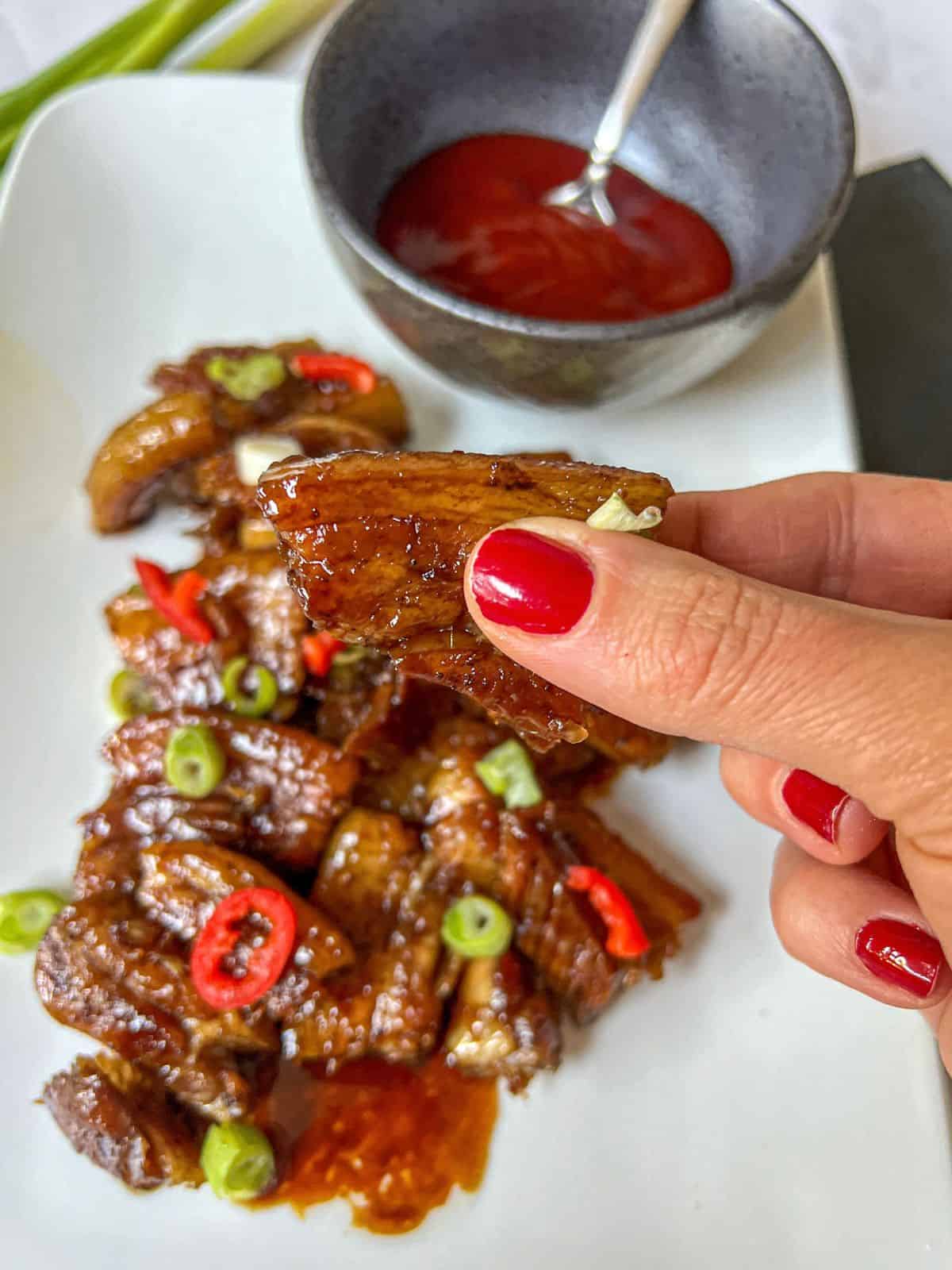 Someone holding up a cooked pork belly slice from a plate of pork belly garnished with chilli and spring onion.