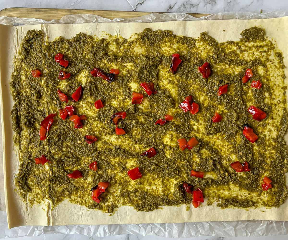 A sheet of uncooked puff pastry with pesto and roasted red peppers on top.
