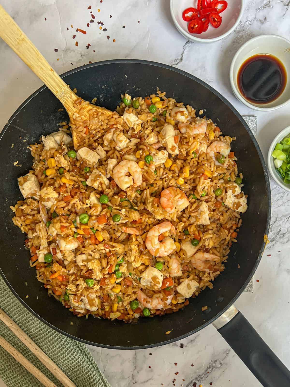 A wok containing chicken & shrimp fried rice with small bowls to the side containing red chilli, soy sauce and spring onion.