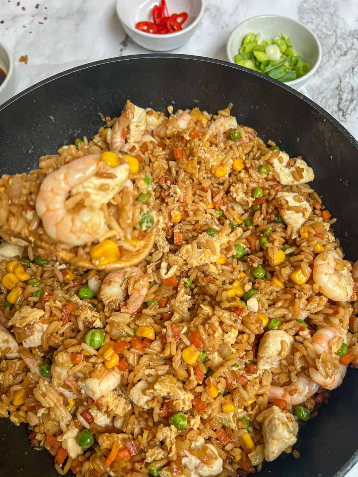Chicken and shrimp fried rice in a wok with little bowls of chilli and spring onion on the side.