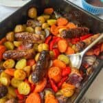 A roasting tin with a spoon in it containing sausages, roasted potatoes, carrots, onions and peppers.