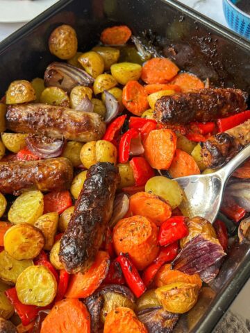 A roasting tin with a spoon in it containing sausages, roasted potatoes, carrots, onions and peppers.